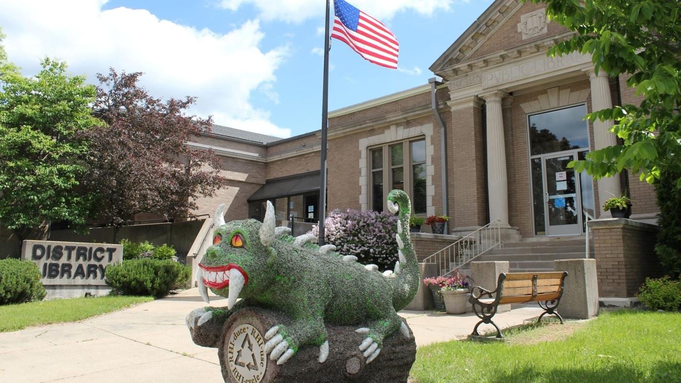 A playful sculpture of a creature in front of a historic building adorned with an American flag in Rhinelander, Wisconsin.