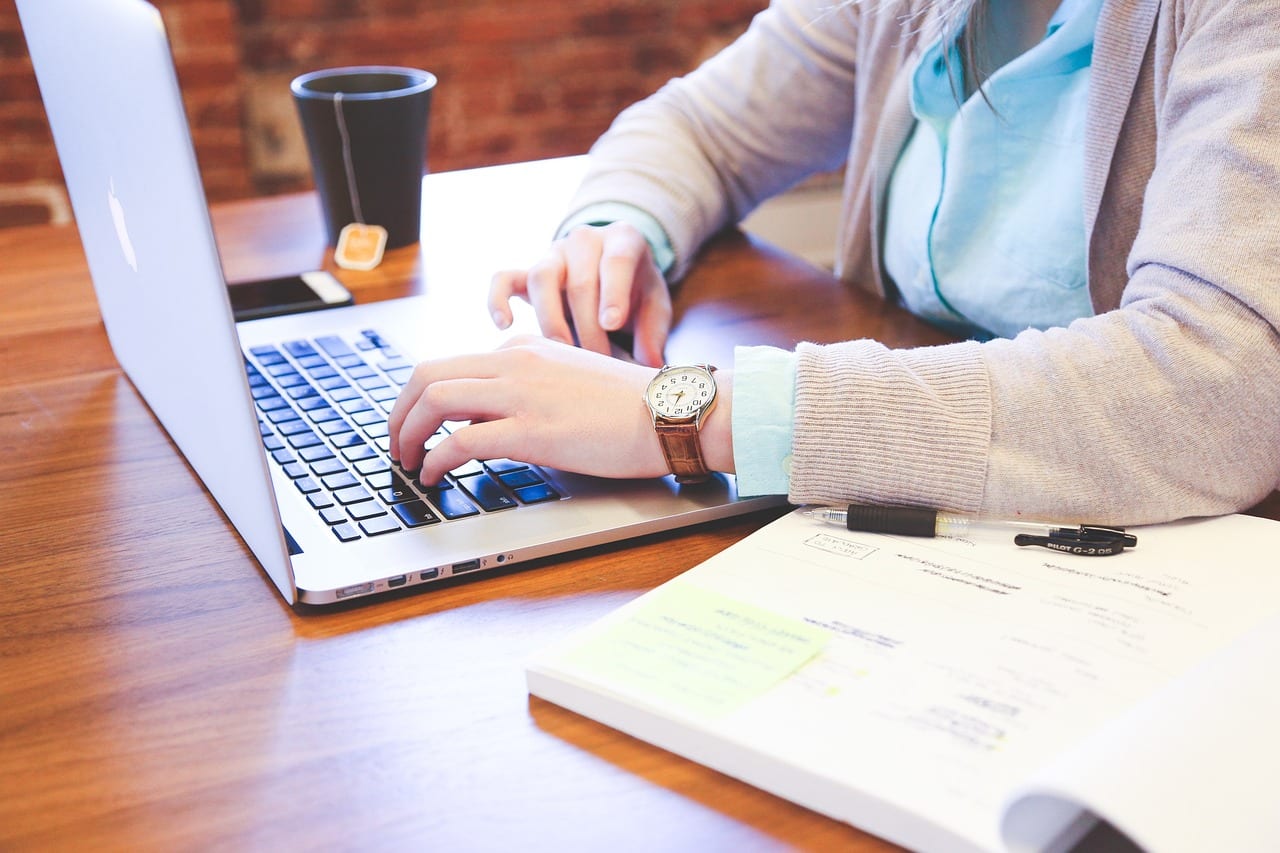 Person typing on a laptop with notebooks and a cup of tea on a wooden desk