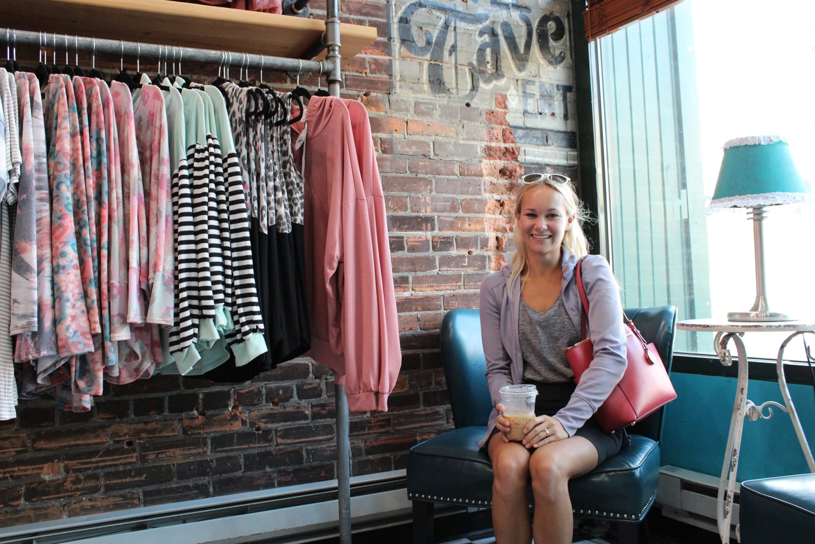 A woman seated inside Brown St. Boutique with clothing racks and a brick wall in the background.