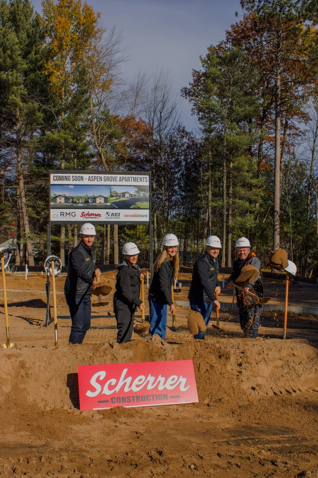 Five people in hard hats turning soil at a construction groundbreaking site in Oneida County, Wisconsin, with trees and a project sign in the background.