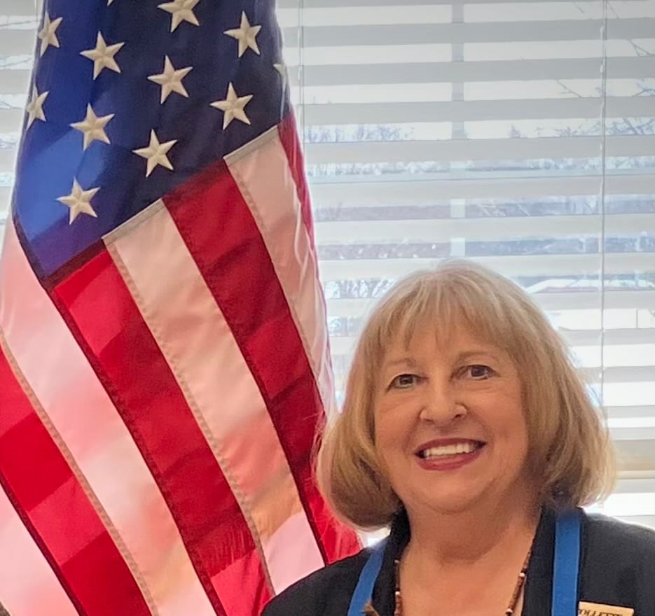 Smiling woman with blonde hair standing in front of an American flag and window blinds, symbolizing leadership in Oneida County, WI