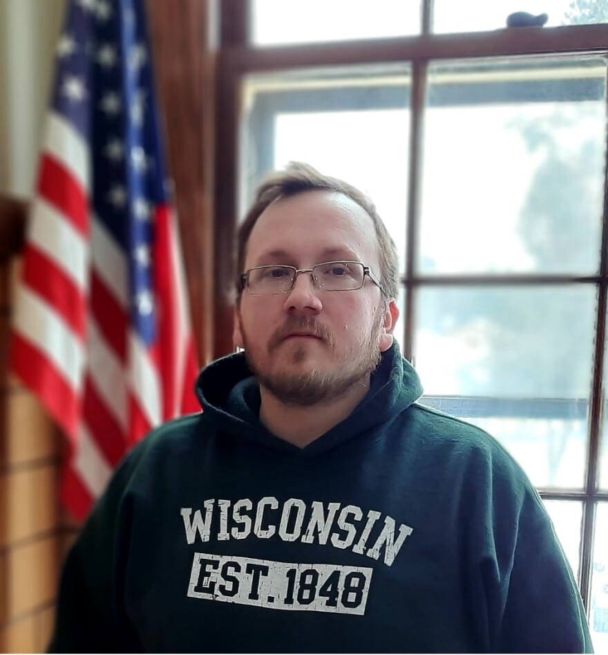 Man in a Wisconsin hoodie standing by a window with an American flag in the background