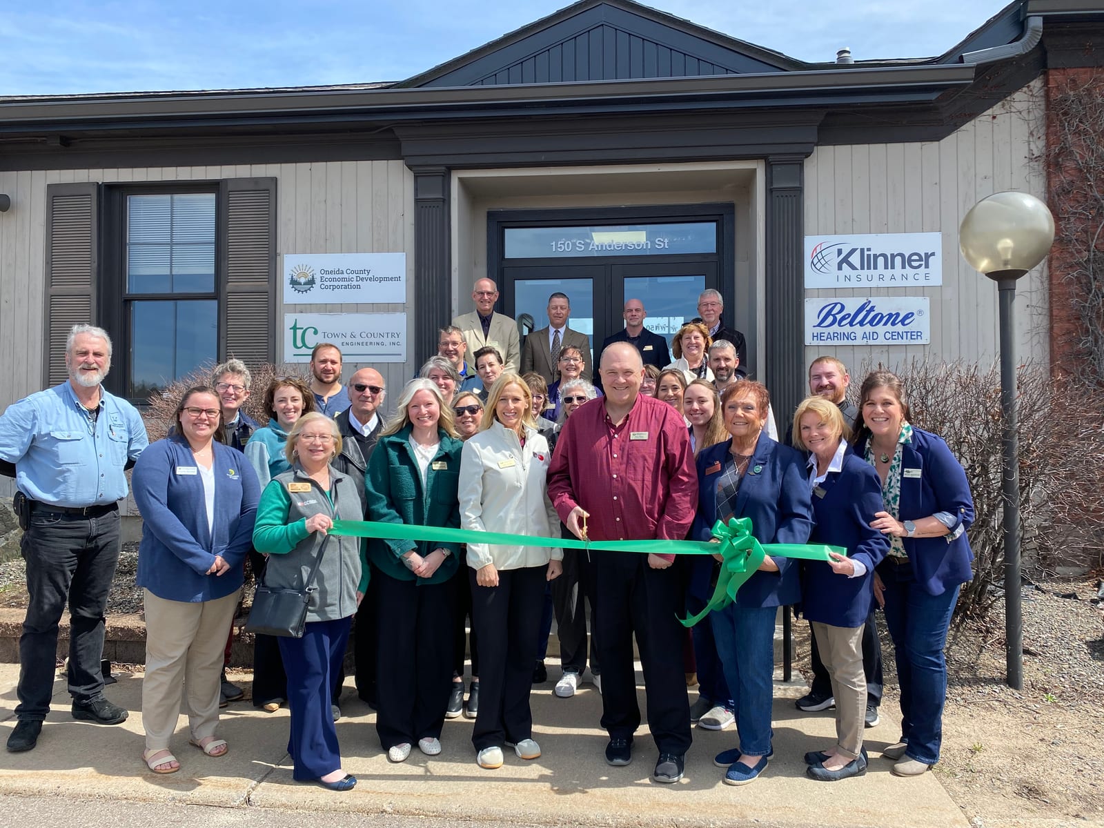 A group of community members and leaders gathered in front of a building, holding a large green ribbon during a ribbon-cutting ceremony for Oneida County Economic Development Corporation.