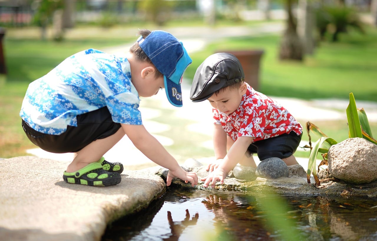 Two children wearing colorful shirt and hats, crouching by a pond in a park