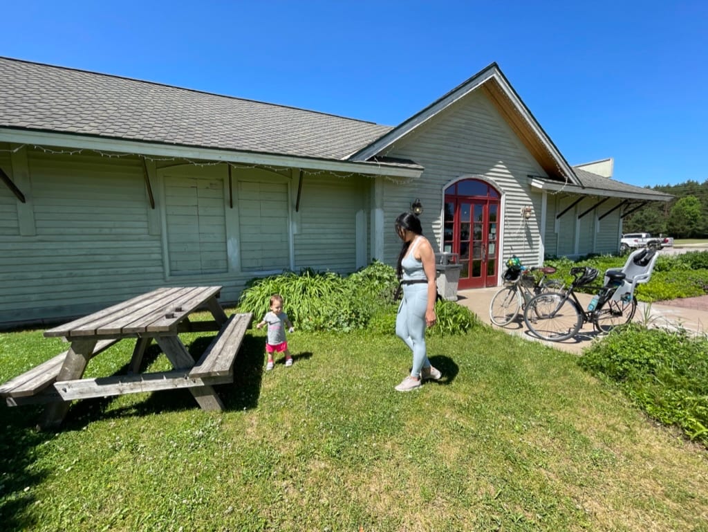 A person walking with a child near a picnic table and bicycles outside a building under clear blue skies