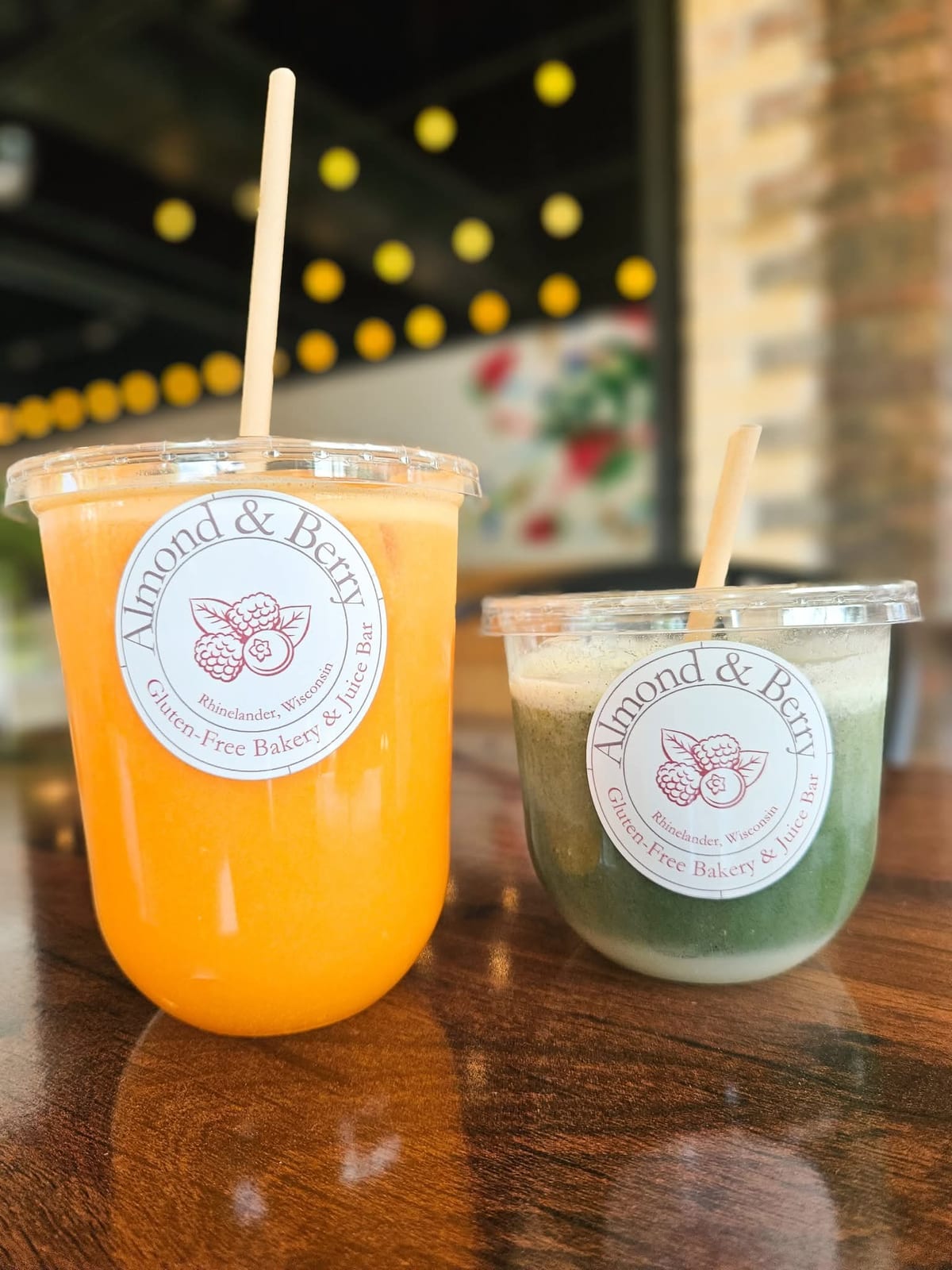 Two clear cups with the 'A&B Juice Bar' label, one filled with a vibrant orange juice and the other with a green juice, both with wooden straws, sitting on a wooden table in a warmly lit café.