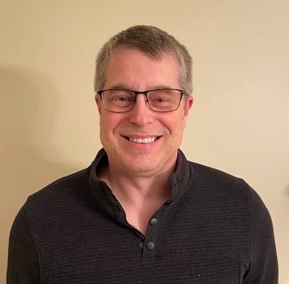 A person with glasses and a dark zip-up shirt, smiling in front of a plain beige wall