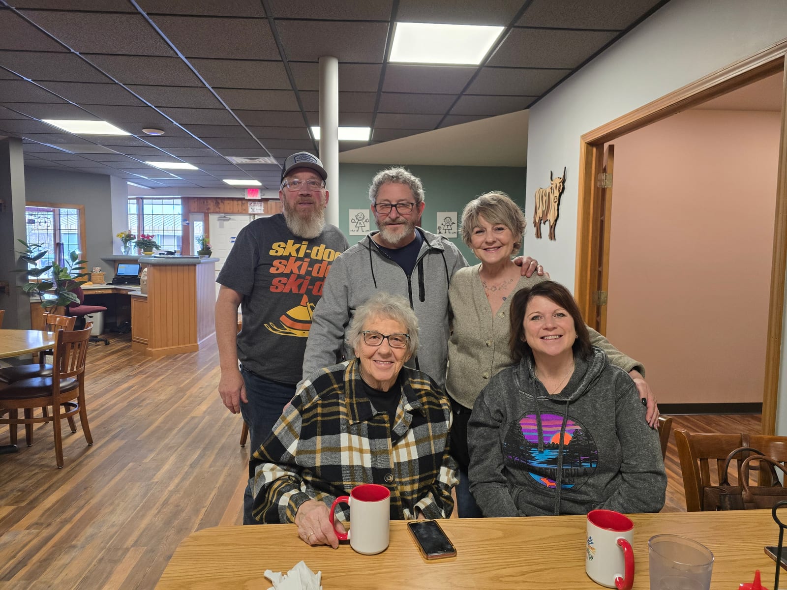 Group of five people sitting and standing around a diner table with coffee mugs in a cozy diner setting.