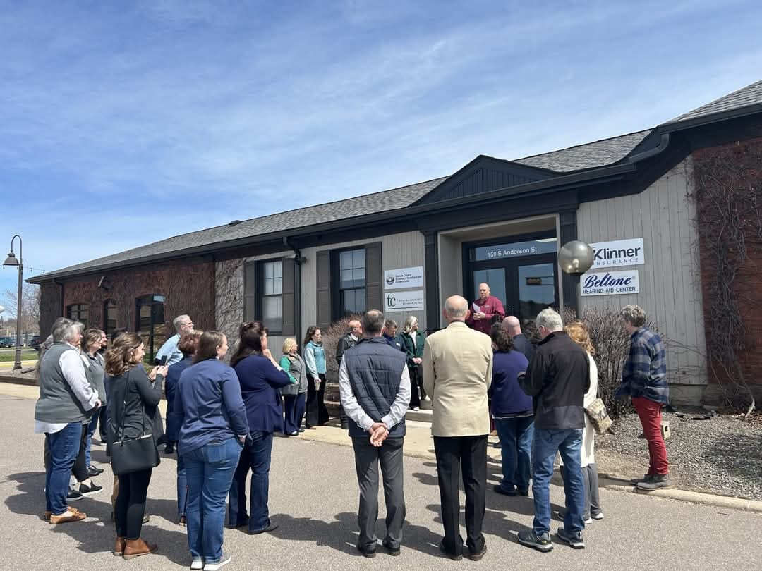 A group of people is gathered in front of a building with blue skies above, attending a ribbon cutting ceremony. The building has signage and a person is speaking on the front steps.