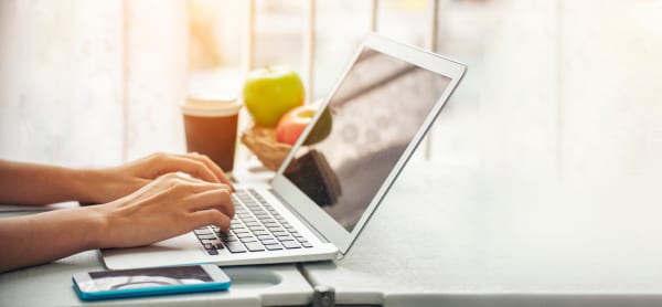 Person typing on a laptop next to a cup of coffee and fruits on a bright morning.