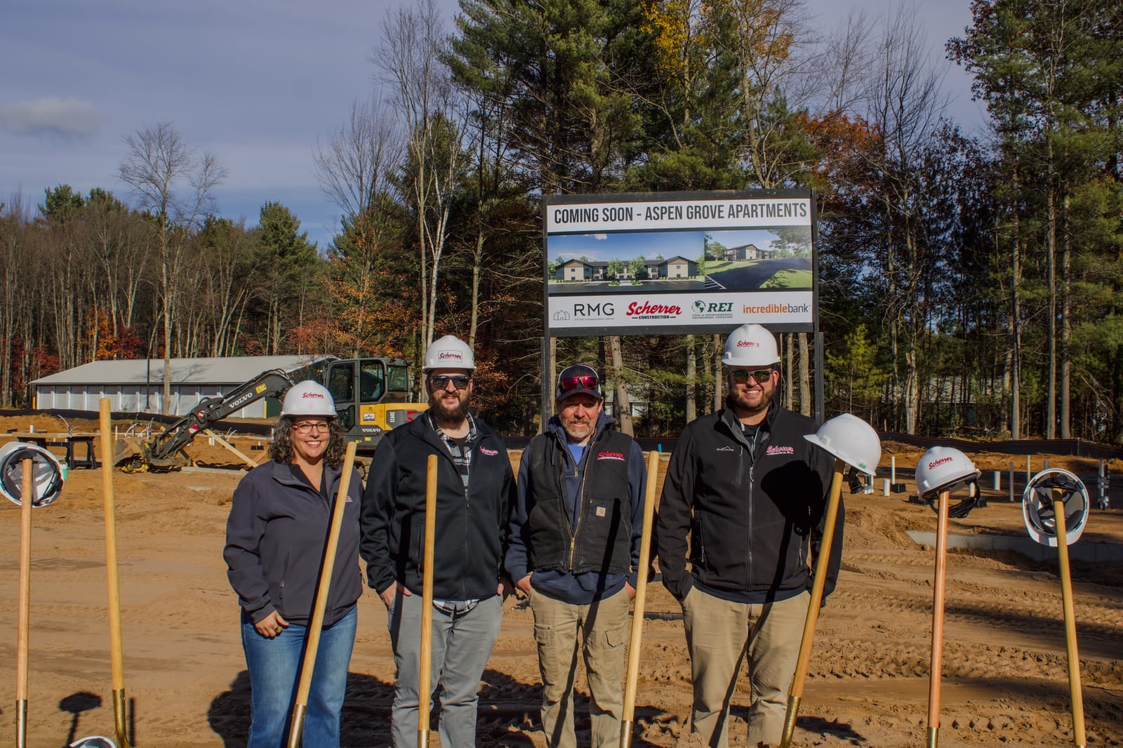 Four people wearing hard hats and holding shovels pose at a construction site in front of a large project sign and surrounded by trees