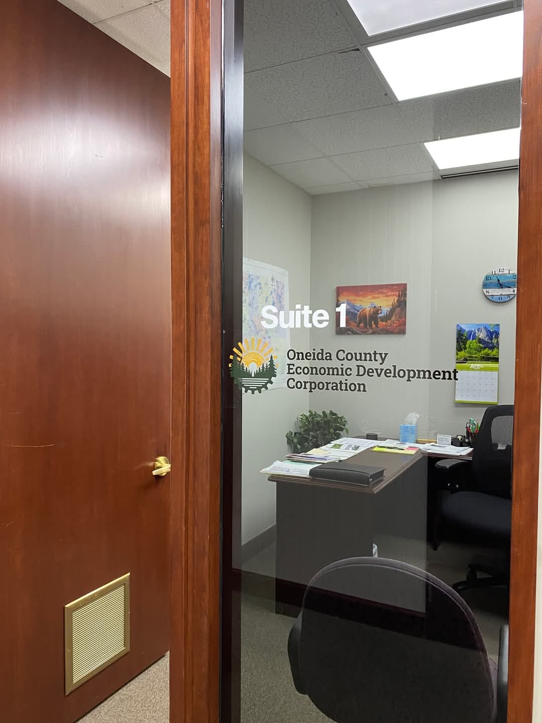 Glass door entry to Oneida County Economic Development Corporation office, showing an interior office with a desk, chair, and wall art in a professional setting