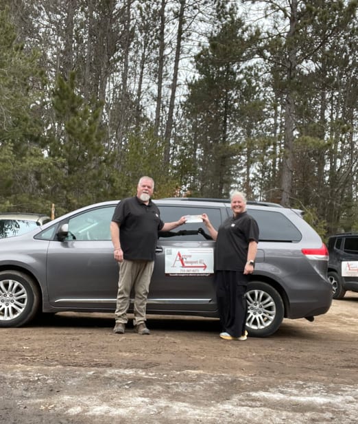 Two people stand by a gray van with Arrow Transport signage, exchanging papers, surrounded by trees in a rural Oneida County setting